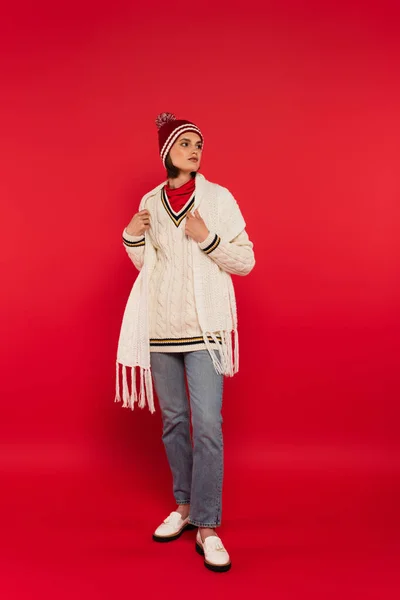 Повна довжина жінки в трикотажному капелюсі і светрі, дивлячись на червоний фон — стокове фото