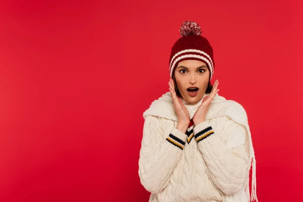 Shocked woman in knitted clothes and hat looking at camera isolated on red — Stockfoto