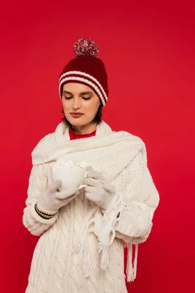 Young woman in warm clothes holding cacao with marshmallows isolated on red — Photo de stock