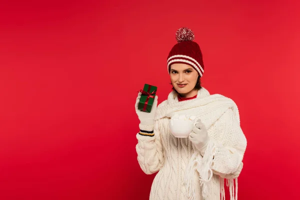 Smiling woman in gloves and warm clothing holding gift and cup with marshmallows isolated on red - foto de stock