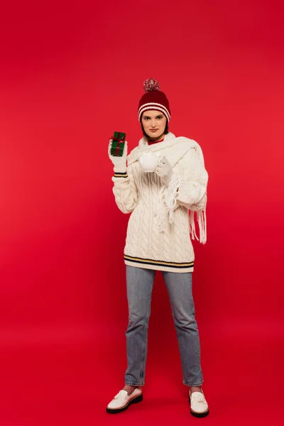 Full length of young woman in warm clothes holding cup with marshmallows and present on red background — Photo de stock