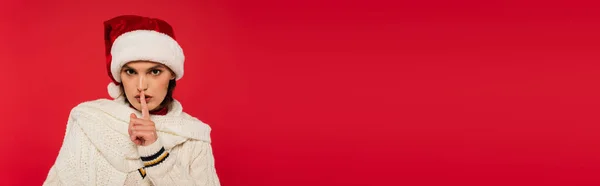 Brunette woman in santa hat showing shh gesture isolated on red, banner — Foto stock