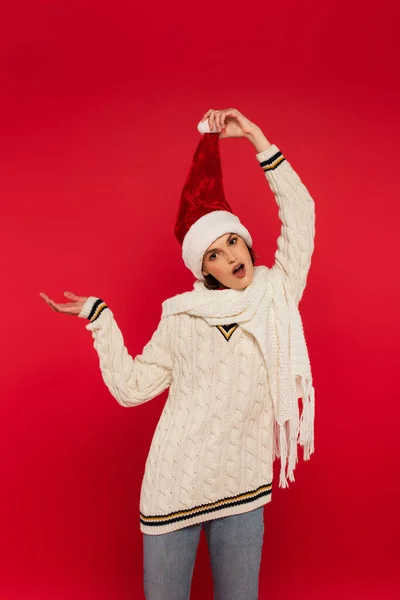 Shocked woman in sweater and santa hat pointing with hand isolated on red — Fotografia de Stock