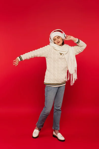 Young woman in santa hat and knitted clothes listening music in headphones on red background — Stockfoto