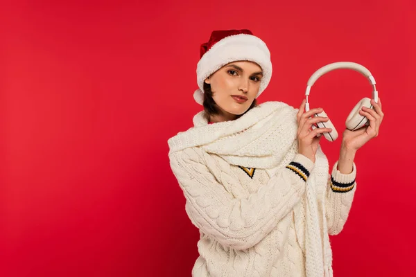 Brunette woman in santa hat holding headphones isolated on red — Stock Photo