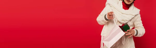 Cropped view of young woman in sweater putting gift box with bow into paper bag on red, banner — Foto stock