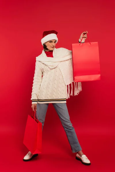 Full length of young woman in santa hat and winter outfit holding shopping bags on red — Stock Photo
