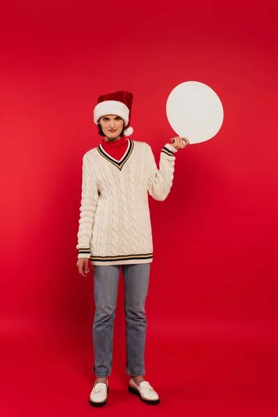 Full length of young woman in santa hat and sweater holding speech bubble on red — Stock Photo