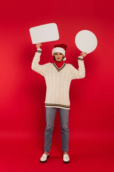 Full length of young woman in santa hat and sweater holding speech bubbles on red — Fotografia de Stock