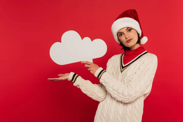 Young woman in santa hat and sweater holding thought bubble and pointing with hand isolated on red — Stock Photo