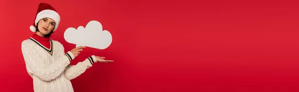 Young woman in santa hat and sweater holding thought bubble and pointing with hand isolated on red, banner — Stock Photo