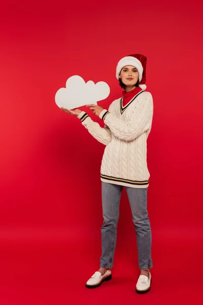 Full length of young woman in santa hat and sweater holding thought bubble on red — Stock Photo
