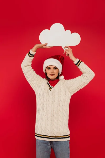 Smiling young woman in santa hat and sweater holding thought bubble above head isolated on red — Stock Photo