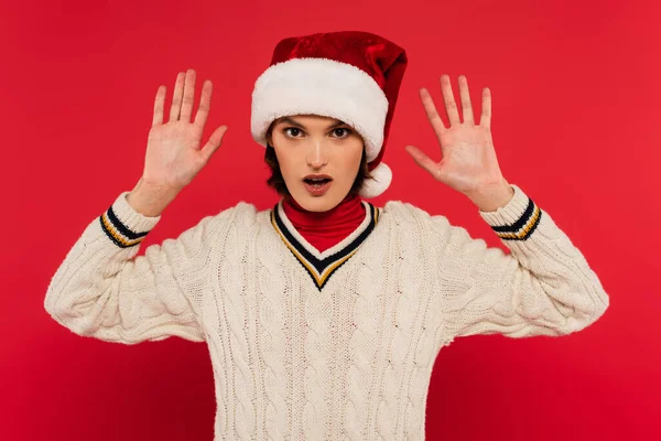 Surprised young woman in santa hat and sweater gesturing and looking at camera isolated on red — Photo de stock