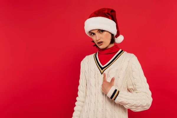 Young woman in santa hat and sweater pointing at herself isolated on red — Photo de stock