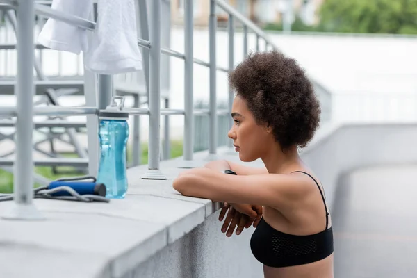 Side view of brunette african american sportswoman with crossed arms near sports bottle and blurred jump rope - foto de stock