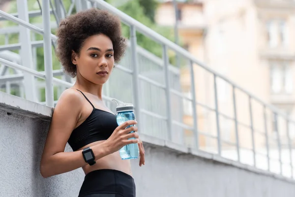 Athletic african american woman in fitness tracker holding sports bottle and looking at camera — Fotografia de Stock