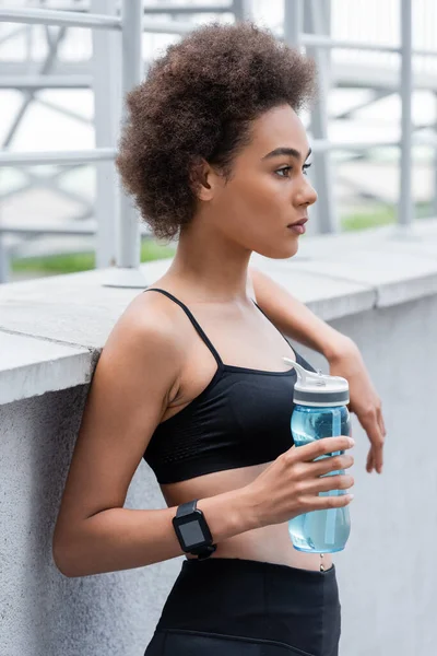 Pretty african american woman in sports bra and fitness tracker holding sports bottle outdoors - foto de stock