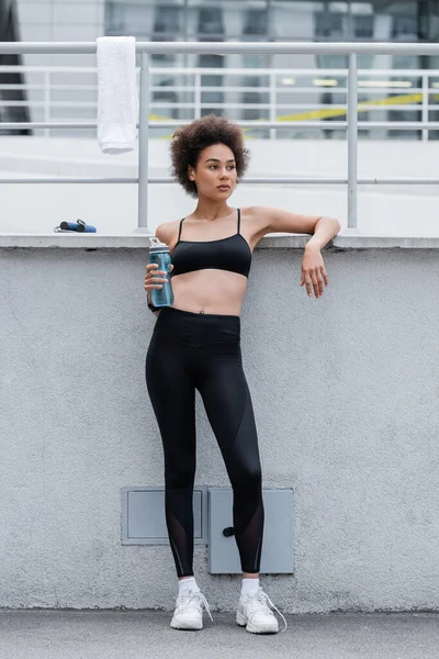 Slim african american woman in black sportswear and white sneakers standing with sports bottle near towel on fence - foto de stock