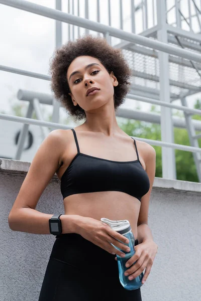 Low angle view of sportive african american woman with sports bottle and fitness tracker looking at camera — Fotografia de Stock