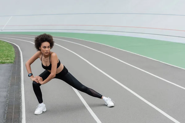 Full length of african american woman in black sportswear doing side lunge exercise while stretching on stadium — Stock Photo