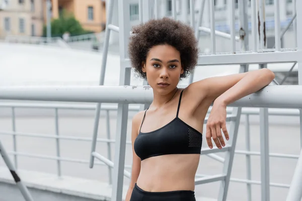 Sportive african american woman in black sports bra looking at camera near fence — Stockfoto