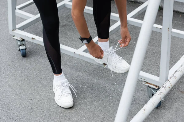 Cropped view of african american woman in fitness tracker tying shoelaces on white sneakers — Foto stock