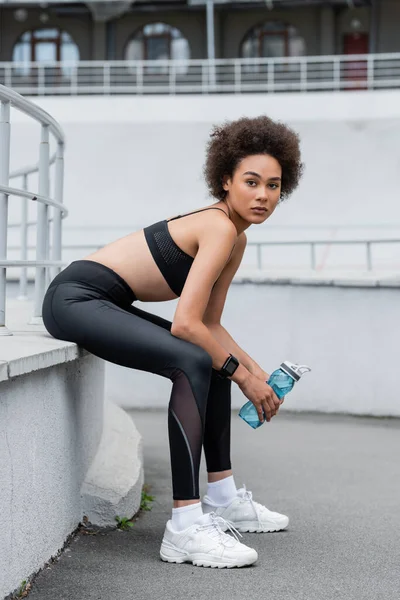 Full length of african american woman in black sportswear and white sneakers sitting with sports bottle — Stockfoto