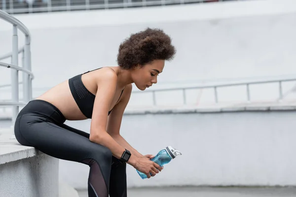 Side view of african american woman in black sportswear sitting with sports bottle on stadium — Fotografia de Stock