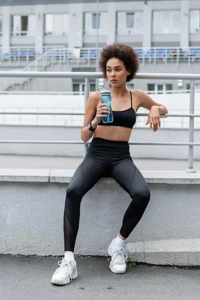 Full length of african american woman in black sportswear and white sneakers sitting with sports bottle — Fotografia de Stock