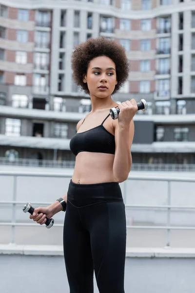 Brunette african american sportswoman working out with dumbbells and looking away — Stockfoto