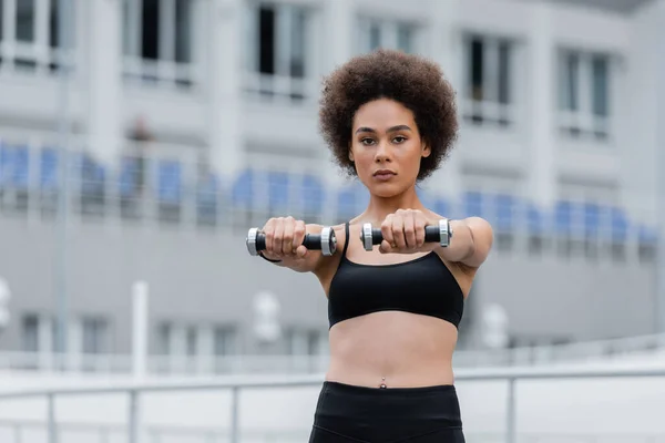 Young african american sportswoman looking at camera while exercising with dumbbells — Photo de stock