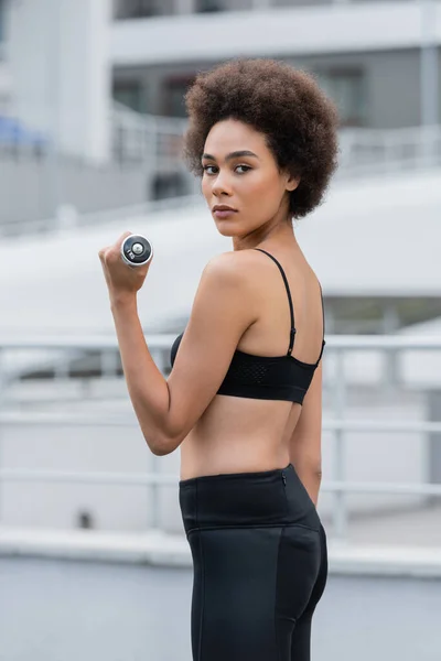 Sportive african american woman in black sportswear training with dumbbell and looking at camera - foto de stock