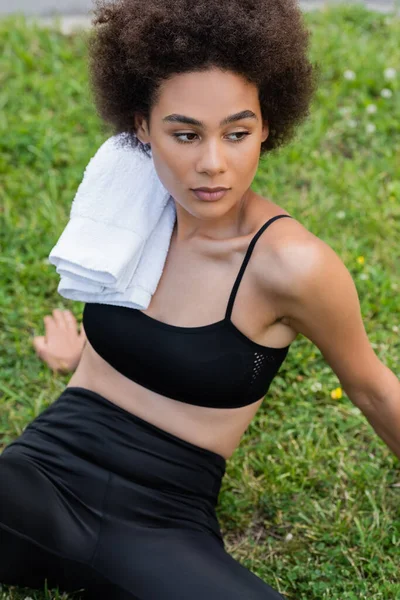 Curly african american woman in black sports bra sitting on lawn with white towel on shoulder — Fotografia de Stock