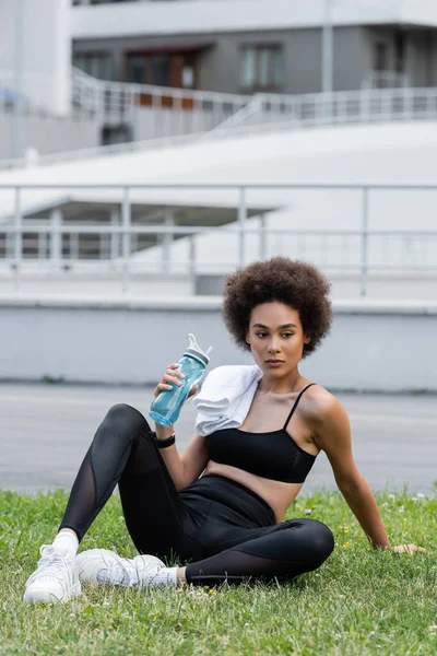 Slender african american woman in black sportswear sitting on lawn with towel and sports bottle — Stock Photo