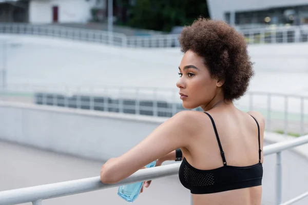 Молодая и кудрявая американка держит спортивную бутылку возле забора — стоковое фото