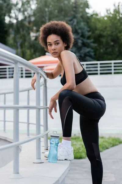 Sportive african american woman stepping on parapet near fence and sports bottle — Foto stock