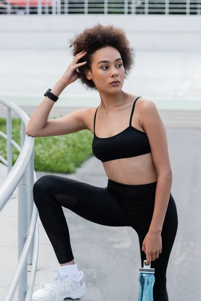 Brunette african american woman in black sportswear holding sports bottle and looking away — Stockfoto