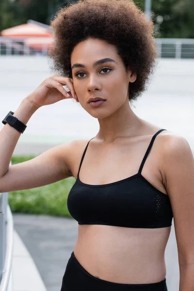 Young african american woman in black sports bra and fitness tracker standing with hand near face — Fotografia de Stock