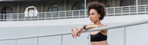 Athletic african american woman in black sports bra leaning on fence and looking away, banner — Stockfoto