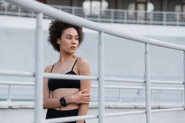 Curly african american woman in black sports bra and fitness tracker standing near fence — Stock Photo