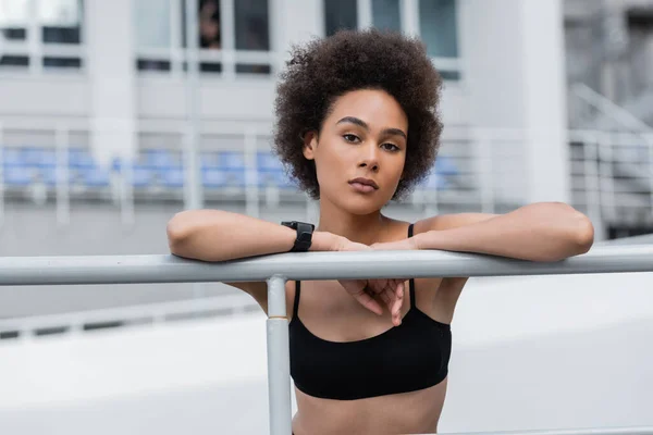 Brunette african american woman with curly hair standing near fence in sports bra — Fotografia de Stock