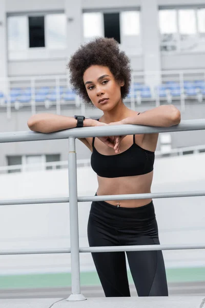 Slender african american woman in sportswear and fitness tracker looking at camera near fence — Stock Photo