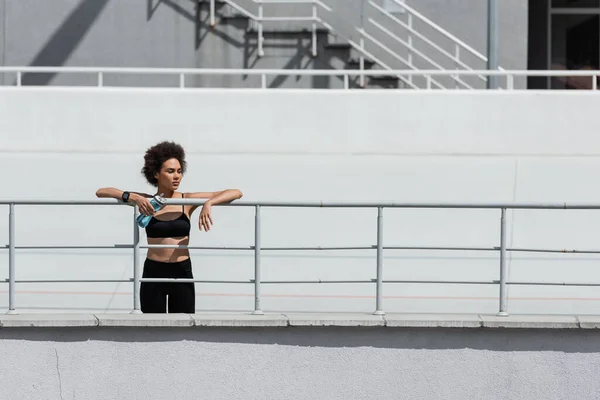 Athletic african american woman in black sportswear standing with sports bottle near fence — Fotografia de Stock