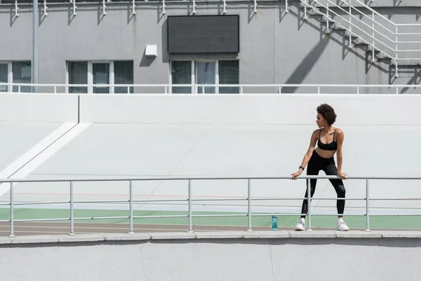 Повна довжина афроамериканської жінки в чорному спортивному одязі, що стоїть біля спортивної пляшки на міському стадіоні — стокове фото