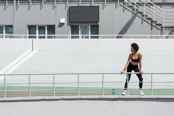 Full length of african american woman in black sportswear standing near sports bottle on city stadium — Foto stock