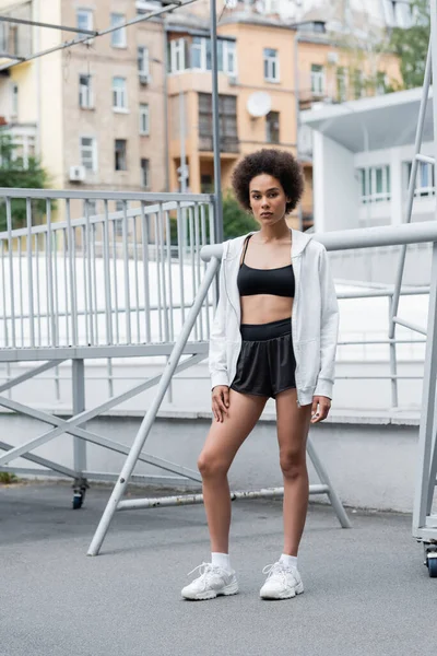 Full length of african american woman in black sportswear and white hoodie standing on city stadium — Stockfoto