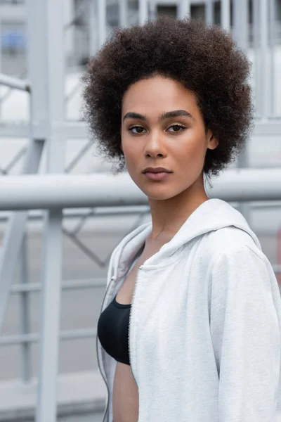 Brunette african american woman in white hoodie looking at camera outdoors - foto de stock