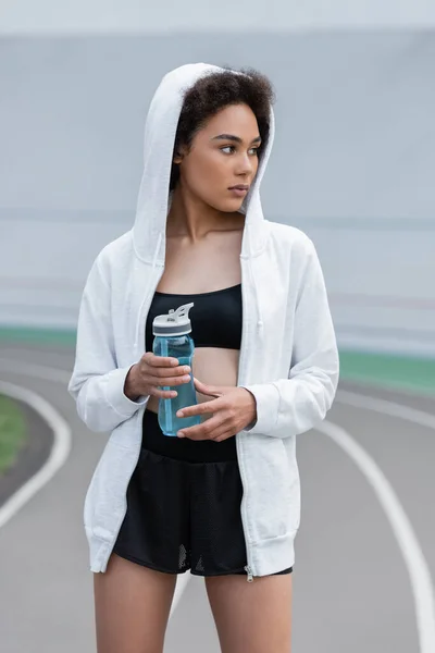 Young african american woman in black sportswear and white hoodie holding sports bottle and looking away — Stock Photo