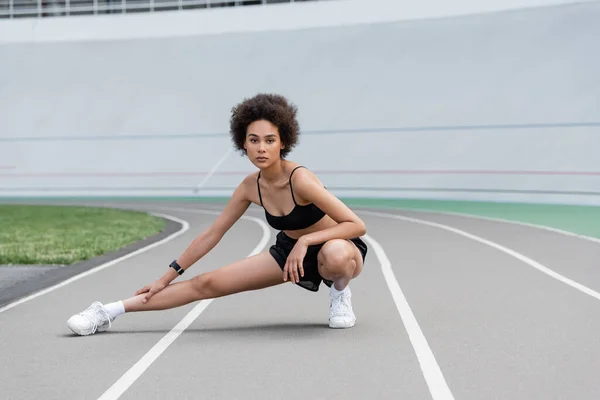 Sportive african american woman stretching leg on stadium and looking at camera - foto de stock